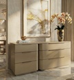 Close-up of a modern, luxury dresser in a smooth champagne finish, featuring a curved silhouette and sleek horizontal handles. The dresser is styled with a gold vase of orchids and decorative books, set against a backdrop of abstract art and marble flooring.