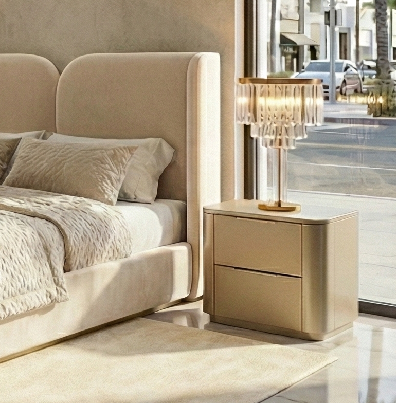 A luxurious contemporary bedroom set in a beige and gold finish, featuring an upholstered bed with textured bedding, two matching nightstands with crystal lamps, and a large dresser with gold artwork above it, all arranged on a marble floor in a showroom with large windows overlooking a palm-lined street.