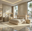 A luxurious contemporary bedroom set in a beige and gold finish, featuring an upholstered bed with textured bedding, two matching nightstands with crystal lamps, and a large dresser with gold artwork above it, all arranged on a marble floor in a showroom with large windows overlooking a palm-lined street.